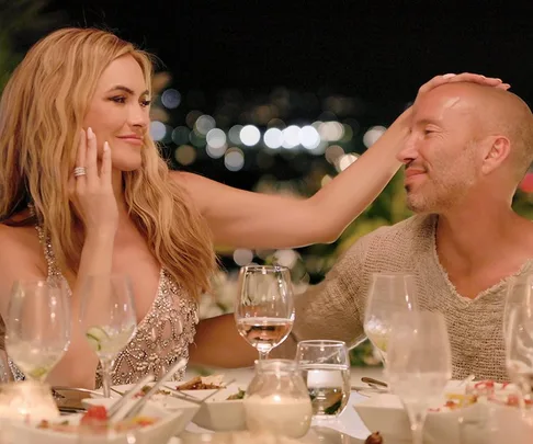 A woman gently touches a man's head at a dinner table, surrounded by dishes and glasses, with city lights in the background.