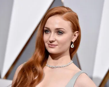 Sophie Turner in a silver dress with long ginger hair at a formal event, wearing earrings and a diamond necklace.