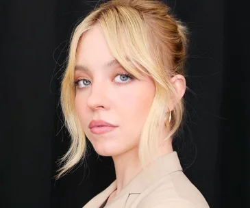 A woman with blonde hair styled in an updo, wearing a beige jacket, against a black background.