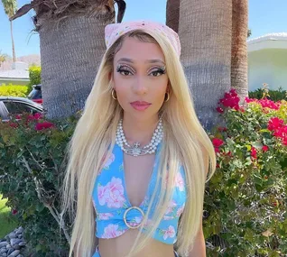 Woman in floral top with a pink bandana and pearl necklaces stands in front of palm trees and pink flowers.