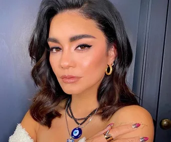 Vanessa Hudgens poses with wavy hair, gold hoop earrings, and layered necklaces against a dark background.