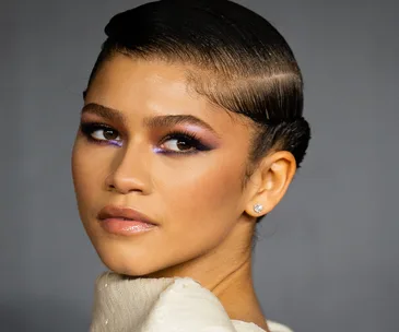 A woman with slicked-back hair, dramatic purple eye makeup, and diamond earrings, wearing a textured white garment.