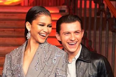 Zendaya and Tom Holland smiling at an event, she wears a sparkling outfit, they stand close against a warm-lit staircase.