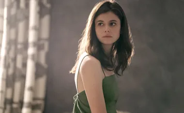 Young woman with long brown hair in a green dress, looking over her shoulder, illuminated by soft lighting indoors.