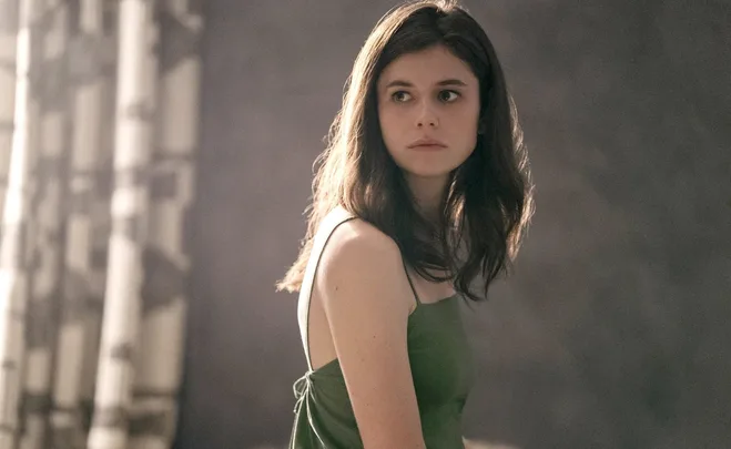 Young woman with long brown hair in a green dress, looking over her shoulder, illuminated by soft lighting indoors.