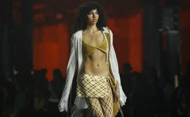 Model in a plaid skirt, gold top, and sheer white shirt walks down a dimly lit runway.