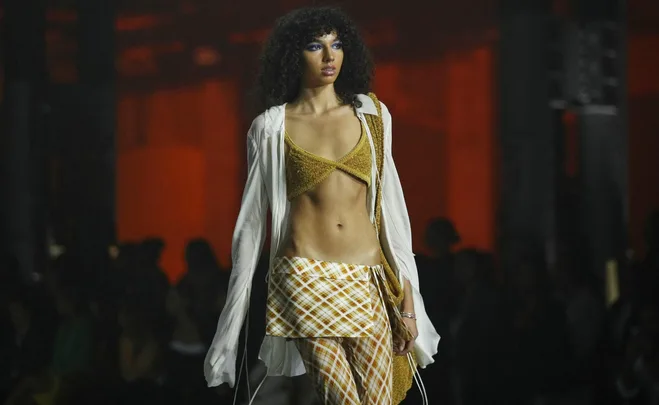 Model in a plaid skirt, gold top, and sheer white shirt walks down a dimly lit runway.