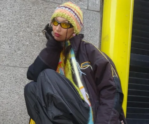 Person in a knit hat, sunglasses, and oversized jacket, sitting near a yellow column and grey wall.