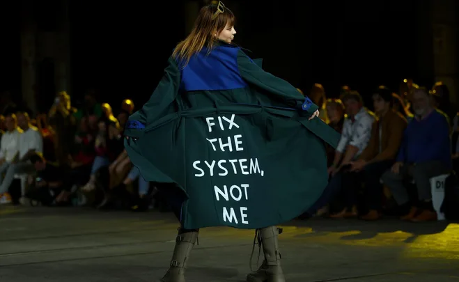 Model on runway in green coat with "Fix the System, Not Me" written on the back, during Australian Fashion Week.