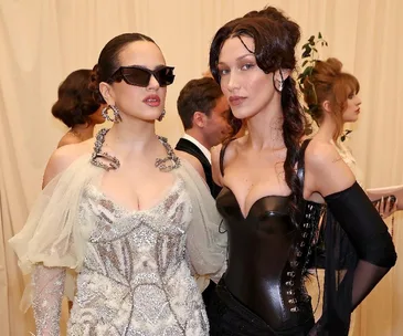 Two women pose in stylish outfits at a formal event. One wears sunglasses and a shimmering dress, the other in a black corset.