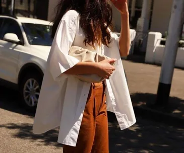 Woman in an oversized white shirt holds a beige clutch on a sunny street with a white SUV in the background.