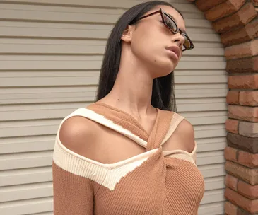 Woman in a stylish beige knit top with sunglasses, standing against a brick arch and shuttered door.