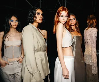 Fashion models backstage, wearing contemporary outfits in neutral tones, with striking eye makeup.