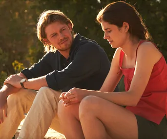 Two people sitting outdoors in the sunlight, engaged in conversation, wearing casual clothing.