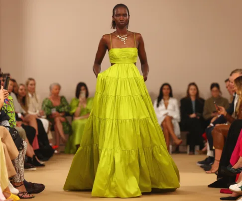 Model wearing a flowing lime green dress walks down a runway with an audience seated on both sides.