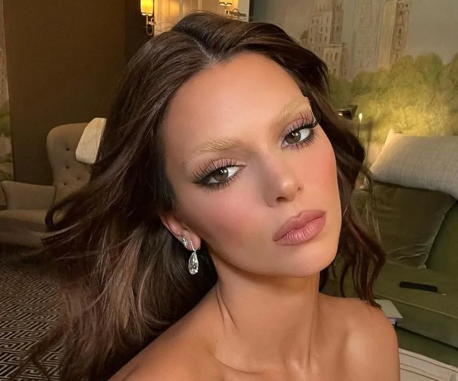 Woman with bleached eyebrows and styled hair, wearing drop earrings in a dimly lit room with elegant decor.