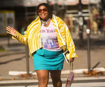 Woman in a yellow striped jacket, blue skirt, and "New York" shirt waves while walking on a sunny street.