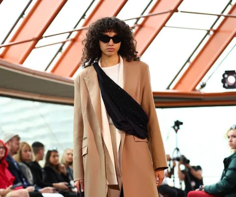 Model in a beige blazer and sunglasses walking on a fashion runway during Australian Fashion Week.