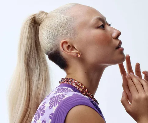 A woman with a platinum blonde ponytail, wearing a purple shirt and jeweled earrings, posed in profile.