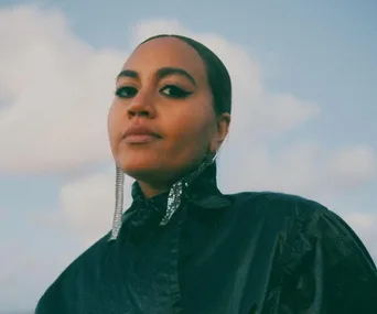 A woman in a shiny black outfit and sparkling earrings gazes confidently under a cloudy sky.