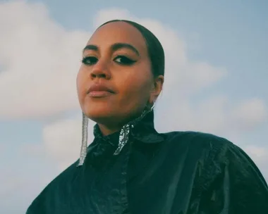 A woman in a shiny black outfit and sparkling earrings gazes confidently under a cloudy sky.