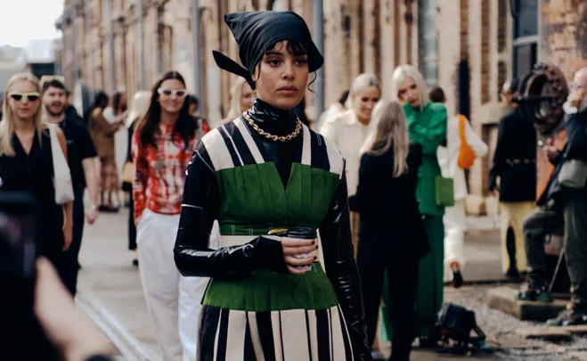 A woman with a headscarf and green dress holds a coffee cup, walking in a stylish crowd on a street.