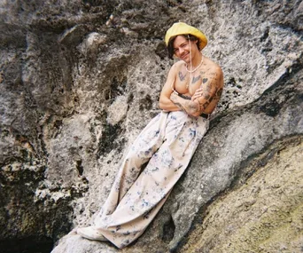 A person wearing a yellow hat and floral pants, sitting on a rocky surface, smiling with arms crossed.