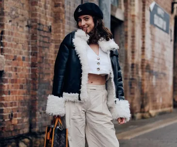 Person in a chic outfit: black jacket with white fur, white crop top, beige pants, black beret, and a designer bag.