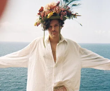 Person wearing a floral crown and white shirt stands with arms outstretched by the ocean.