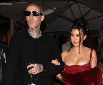 A man in dark sunglasses and tattoos holds a woman's arm; she's in a red dress at an evening event.