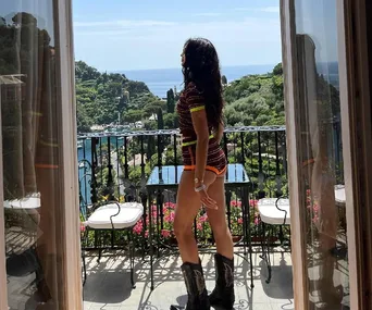 Woman in a striped outfit and boots stands on a balcony overlooking a scenic coastal view.