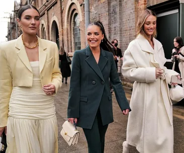 Three women in stylish layered outfits walking outdoors, smiling and carrying handbags.