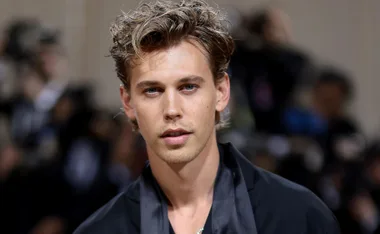 A person with light hair wearing a dark shirt at a formal event, with a blurred background of people and lights.