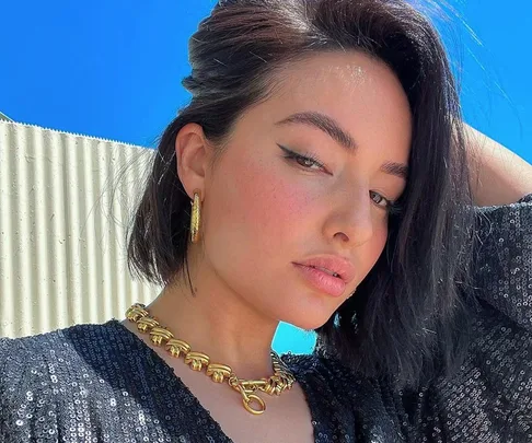 Woman with winged eyeliner and blush poses, wearing gold jewelry and a sparkly black top against a bright sky.