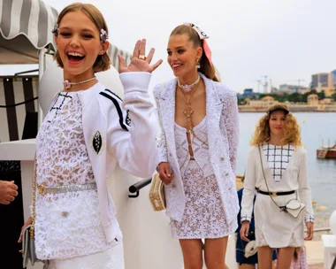 Models in white lace outfits walk up stairs at Chanel Cruise 2023, smiling by a marina backdrop.