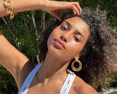 Woman with curly hair, wearing large gold earrings and a bracelet, in a white top, posing outdoors in sunlight.