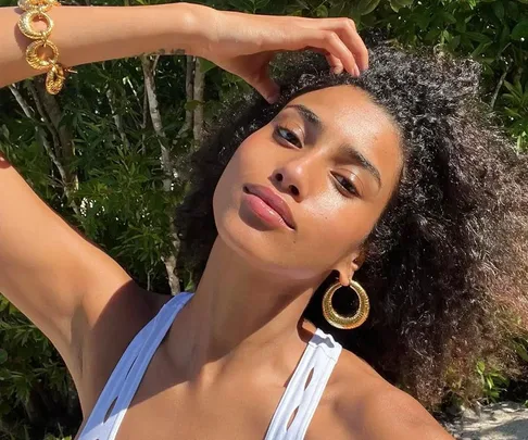 Woman with curly hair, wearing large gold earrings and a bracelet, in a white top, posing outdoors in sunlight.