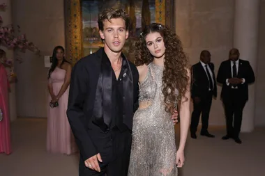 A man and a woman in formal attire pose together at a gala event with people in the background.