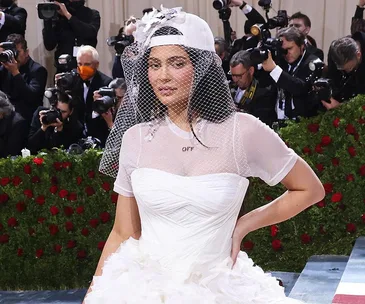 A person in a white gown and cap with a veil stands on a red carpet, surrounded by photographers.