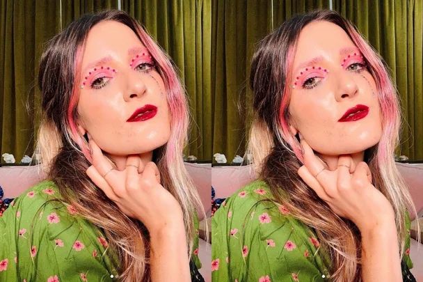 Woman with pink eye makeup and shiny red lips, wearing a green floral blouse, posing for the camera.