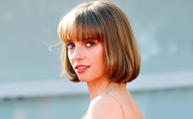 A person with short brown hair, wearing a strapless outfit, looks over their shoulder outdoors.