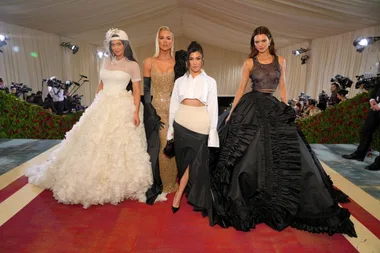 Four women pose in elegant gowns at the Met Gala, surrounded by cameras and a decorated venue.