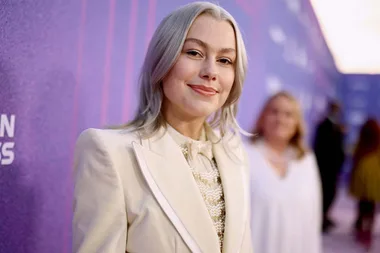 A person with silver hair in a cream outfit smiles in front of a purple background.