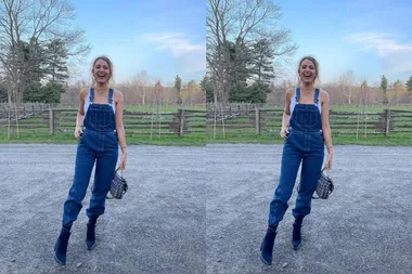 A person wearing denim overalls and black boots, standing outdoors with a wooden fence and trees in the background.