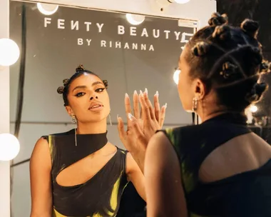 Woman with Bantu knots applying makeup in front of a mirror labeled "Fenty Beauty by Rihanna."