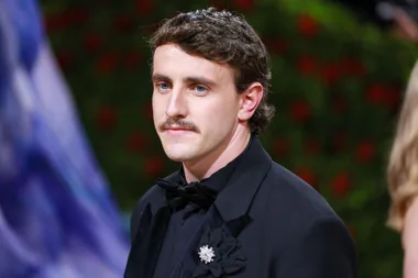 A person in formal attire with a mustache, wearing a black suit and bowtie, stands in front of a green leafy backdrop.