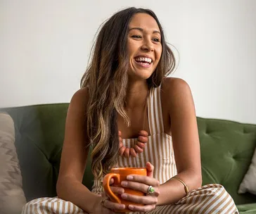 Smiling woman holding a mug, seated on a green couch, wearing a striped outfit and necklace.