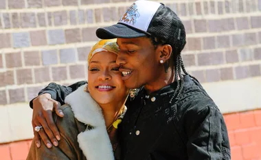 Two individuals embrace and smile against a brick wall backdrop.