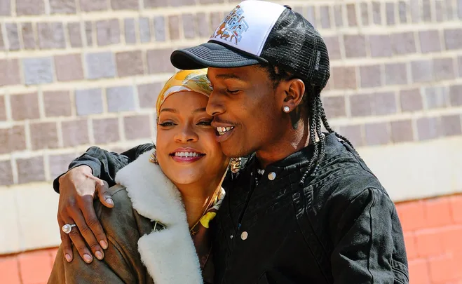 Two individuals embrace and smile against a brick wall backdrop.