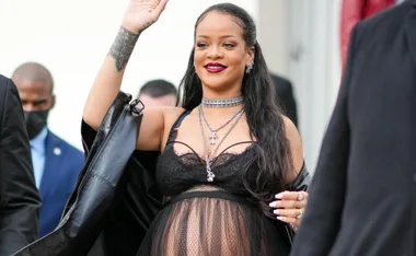 Pregnant woman waves, wearing a sheer black outfit with jewelry, flanked by people in suits.